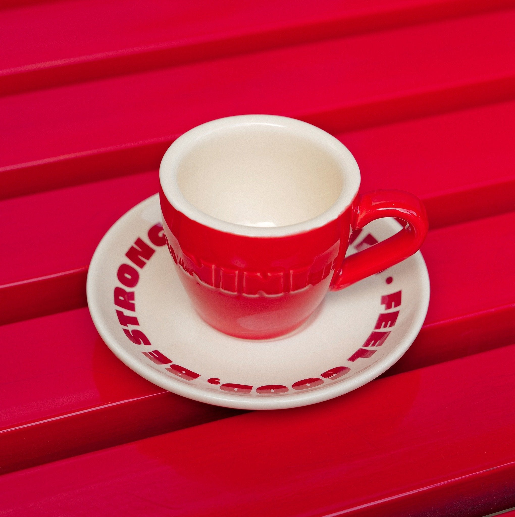 Rote Café NINI Keramik Espressotasse mit cremefarbener Innenseite und eingeprägtem NINI Logo auf cremefarbener Untertasse mit rotem „Feel Good Be Strong" Schriftzug, Produktbild auf rotem Lamellenuntergrund