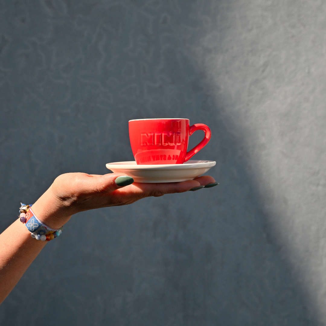 Rote Café NINI Keramik Espressotasse mit weißer Untertasse und eingeprägtem NINI Logo wird auf einer Hand präsentiert, Einzelproduktbild vor grauem Hintergrund