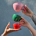 Drei Café NINI Keramik Tassen für Cappuccino, Kaffee und Espresso in Pink, Grün und Rot werden von Händen in die Höhe gehalten, Hochformat Produktbild mit eingeprägtem NINI Logo