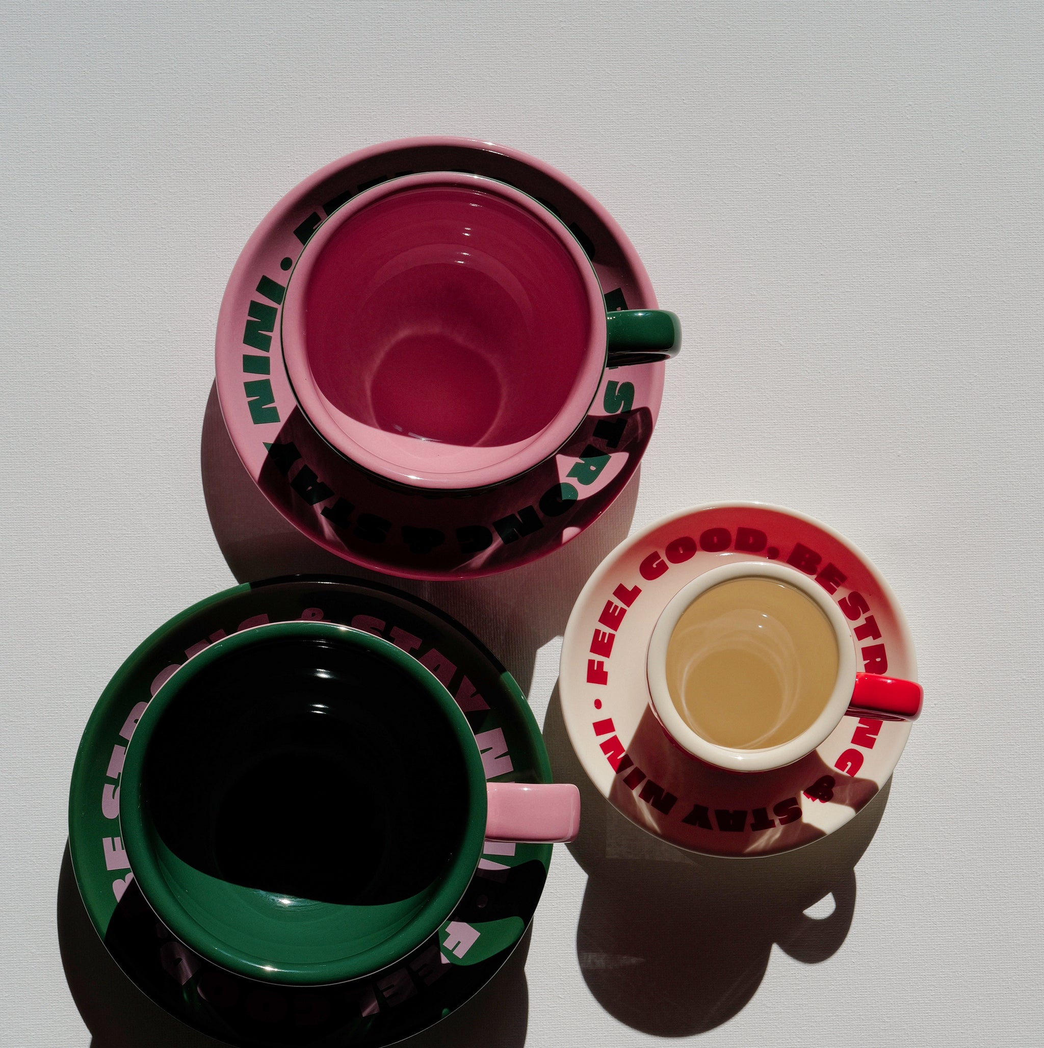 Draufsicht auf drei Café NINI Keramik Tassen in Pink, Grün und Rot mit passenden Untertassen und „Feel Good Be Strong Stay NINI" Schriftzug, eine Tasse mit Kaffee gefüllt, Produktbild auf weißem Hintergrund