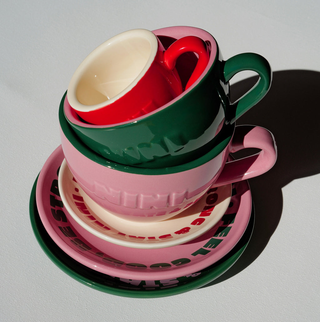 Drei gestapelte Café NINI Keramik Tassen in Pink, Grün und Rot mit passenden Untertassen und eingeprägtem NINI Logo, Produktbild von oben auf weißem Hintergrund
