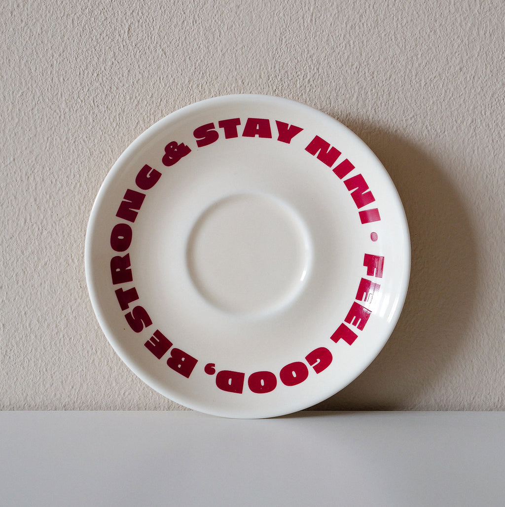 Cremefarbene Café NINI Keramik Untertasse mit rotem „Feel Good Be Strong Stay NINI" Schriftzug, frontale Einzelansicht vor weißem Hintergrund