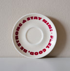 Cremefarbene Café NINI Keramik Untertasse mit rotem „Feel Good Be Strong Stay NINI" Schriftzug, frontale Einzelansicht vor weißem Hintergrund