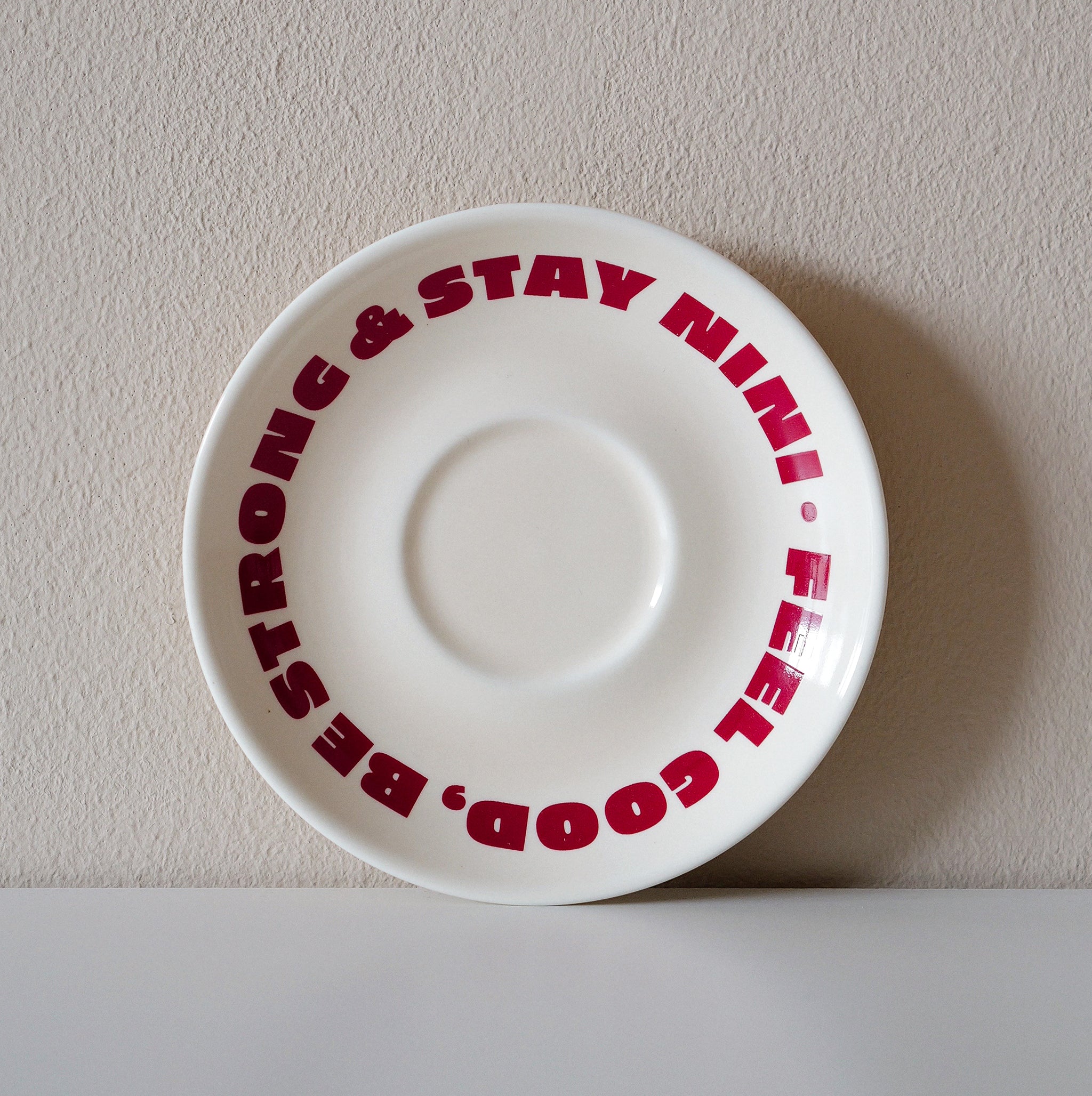 Cremefarbene Café NINI Keramik Untertasse mit rotem „Feel Good Be Strong Stay NINI" Schriftzug, frontale Einzelansicht vor weißem Hintergrund