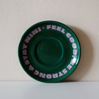 Grüne Café NINI Keramik Untertasse mit pinkem „Feel Good Be Strong Stay NINI" Schriftzug, frontale Einzelansicht vor weißem Hintergrund