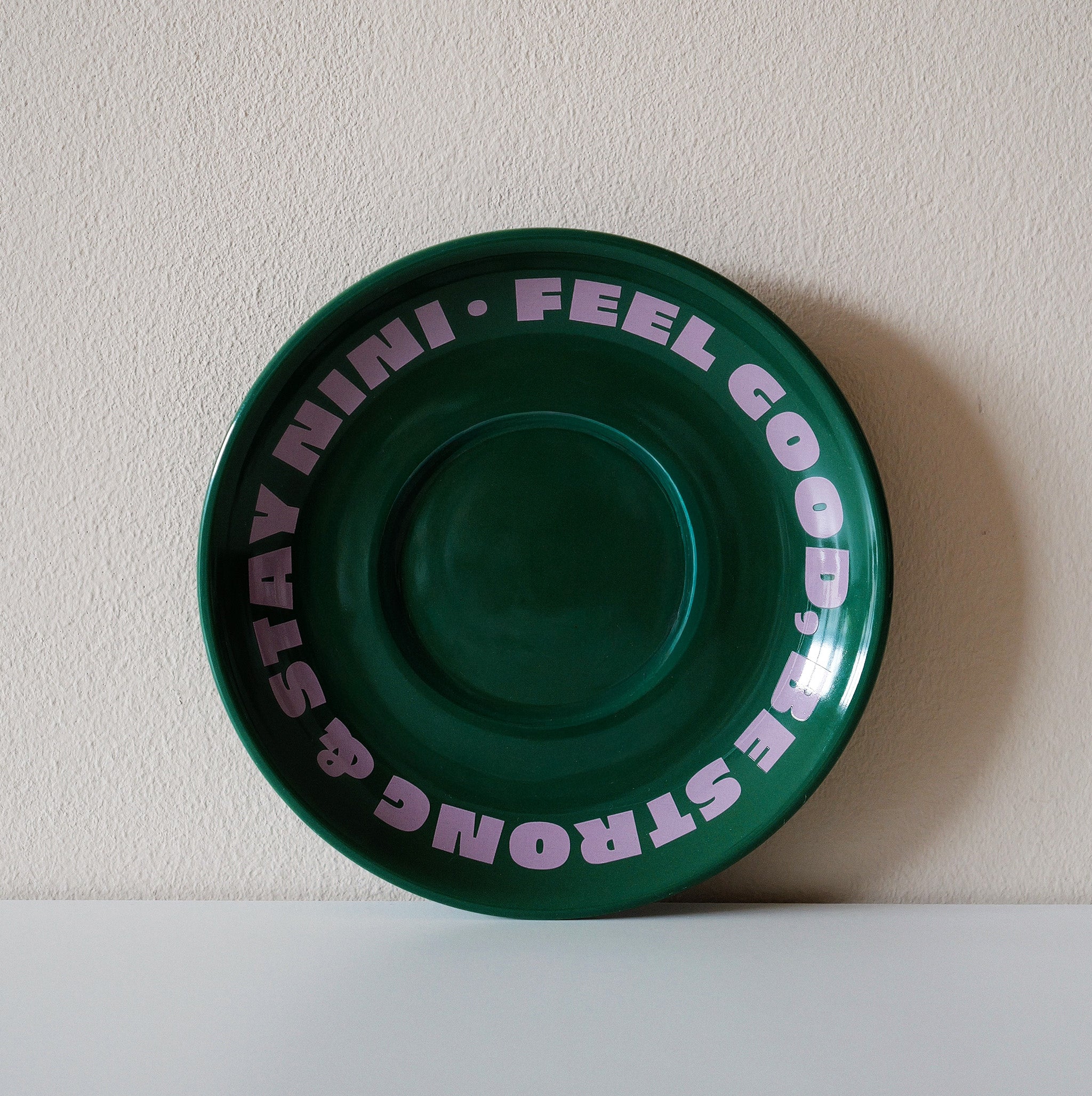 Grüne Café NINI Keramik Untertasse mit pinkem „Feel Good Be Strong Stay NINI" Schriftzug, frontale Einzelansicht vor weißem Hintergrund