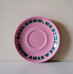 Pinke Café NINI Keramik Untertasse mit grünem „Feel Good Be Strong Stay NINI" Schriftzug, frontale Einzelansicht vor weißem Hintergrund