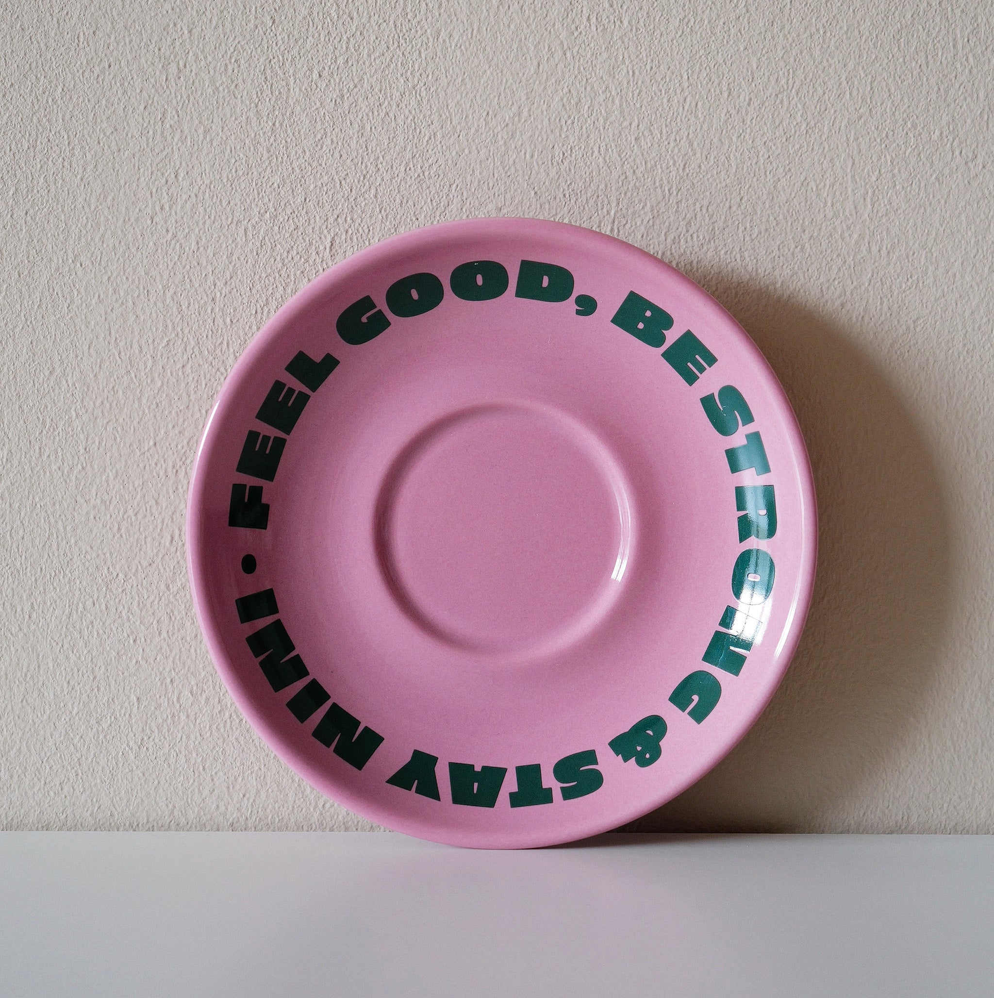 Pinke Café NINI Keramik Untertasse mit grünem „Feel Good Be Strong Stay NINI" Schriftzug, frontale Einzelansicht vor weißem Hintergrund