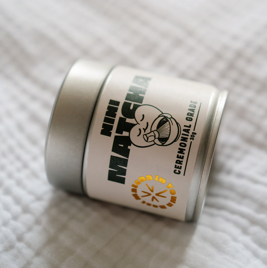 Nahaufnahme der silbernen NINI Matcha Dose Ceremonial Grade 30g liegend auf weißem Stoff, detailliertes Etikett mit NINI Matcha Logo und goldenem „Matcha in Town" Stempel sichtbar