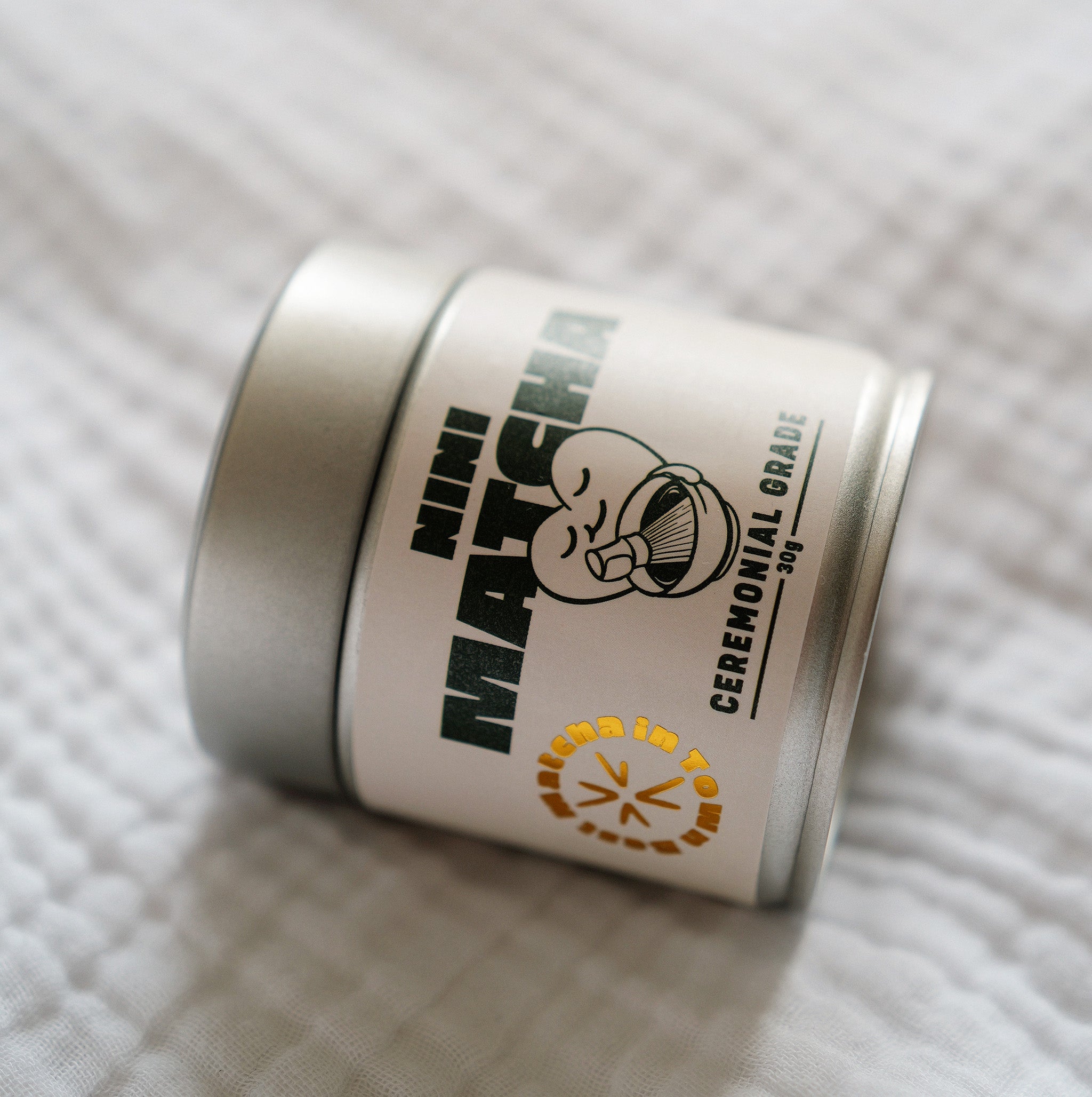 Nahaufnahme der silbernen NINI Matcha Dose Ceremonial Grade 30g liegend auf weißem Stoff, detailliertes Etikett mit NINI Matcha Logo und goldenem „Matcha in Town" Stempel sichtbar