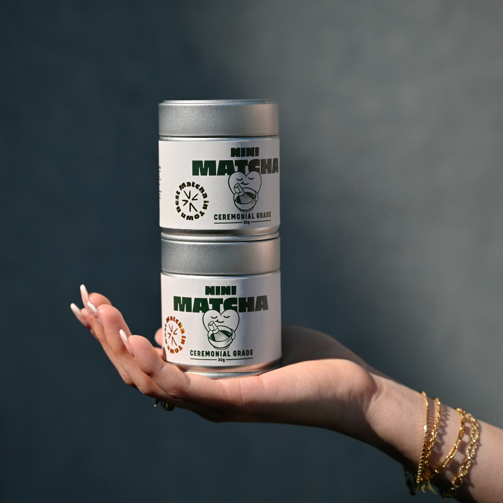 Zwei gestapelte silberne NINI Matcha Dosen Ceremonial Grade 30g werden auf einer Hand balanciert, Produktbild vor dunkelblauem Hintergrund mit sichtbarem NINI Matcha Etikett
