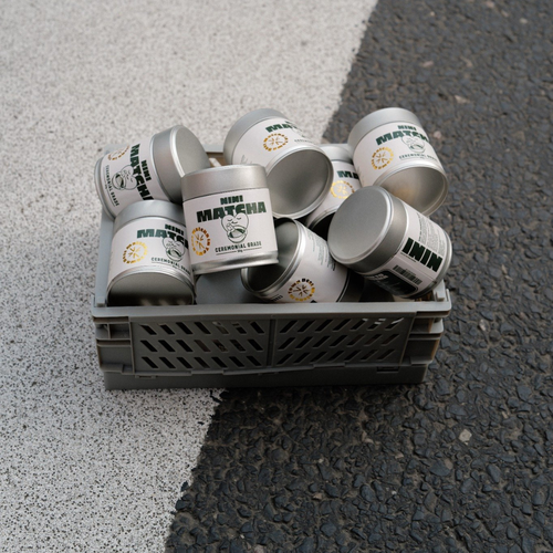 Mehrere silberne NINI Matcha Dosen Ceremonial Grade 30g liegen in einer grauen Transportkiste auf Asphalt, Mengenproduktbild von oben mit sichtbarem NINI Matcha Etikett