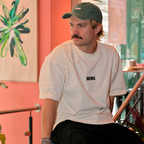 Mann sitzt im Café NINI und trägt weißes Oversized T-Shirt mit schwarzem NINI Frontprint und grüne NINI Cap, lässiges Herren Lookbook Instore