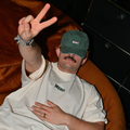 Mann liegt entspannt auf einem Sofa und trägt weißes Café NINI Oversized T-Shirt mit grünem NINI Frontprint und grüne NINI Cap, zeigt Peace Zeichen, lässiges Indoor Lifestyle Lookbook