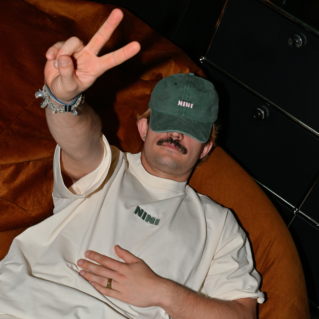 Mann liegt entspannt auf einem Sofa und trägt weißes Café NINI Oversized T-Shirt mit grünem NINI Frontprint und grüne NINI Cap, zeigt Peace Zeichen, lässiges Indoor Lifestyle Lookbook