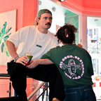 Mann und Frau im Café NINI, Mann trägt weißes Oversized T-Shirt mit NINI Frontprint und grüne Cap, hält grünen Matcha Drink, Frau trägt grünes Café NINI Oversized T-Shirt mit pinkem „Feel Good Be Strong Stay NINI" Backprint