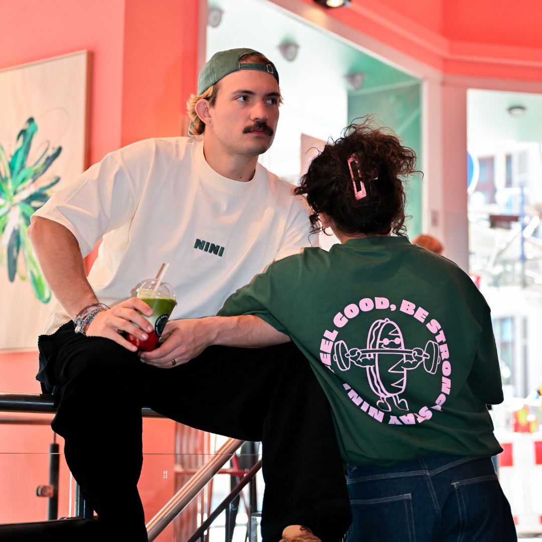 Mann und Frau im Café NINI, Mann trägt weißes Oversized T-Shirt mit NINI Frontprint und grüne Cap, hält grünen Matcha Drink, Frau trägt grünes Café NINI Oversized T-Shirt mit pinkem „Feel Good Be Strong Stay NINI" Backprint